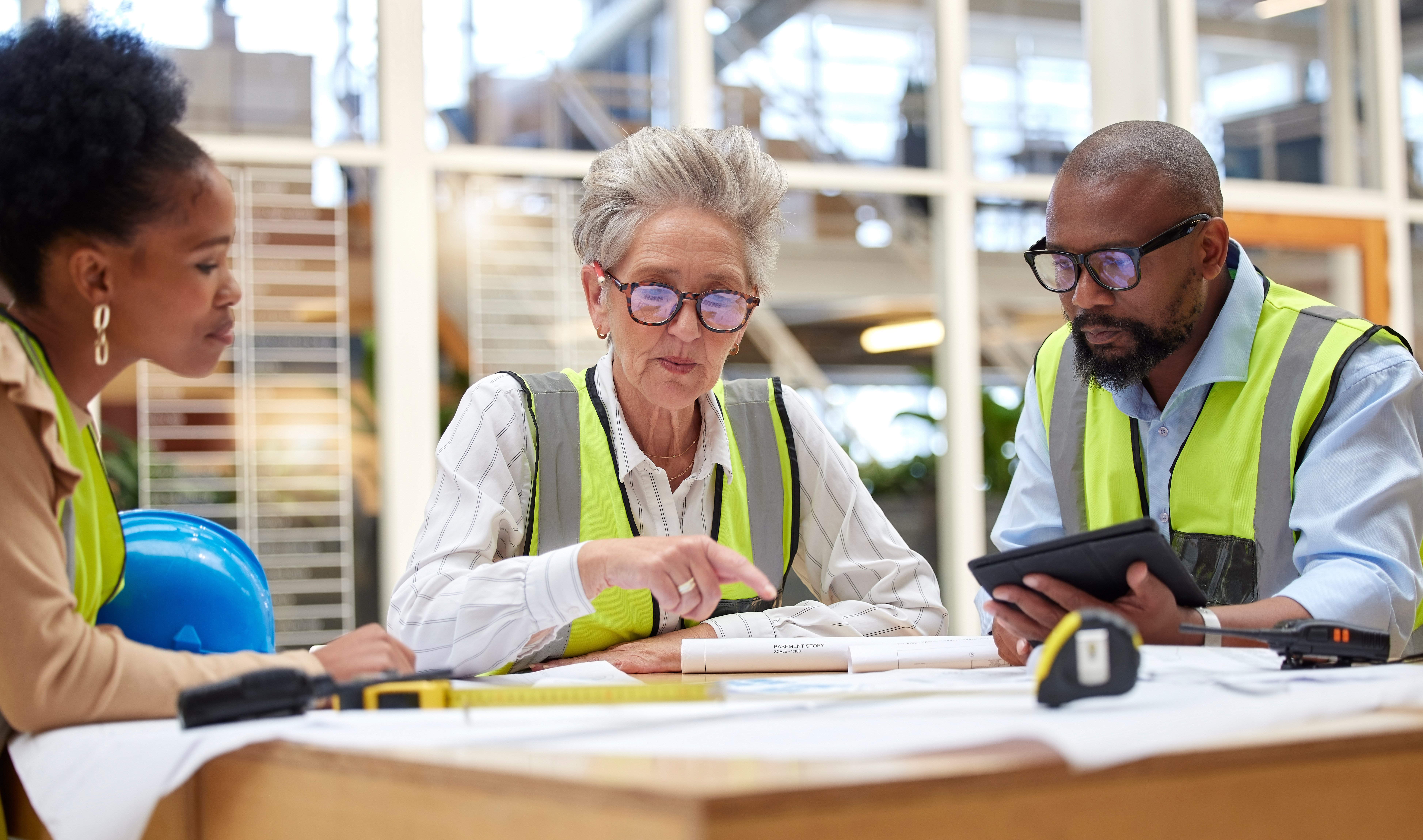 Three workers of varying ages in work vests looking at papers on a table. 