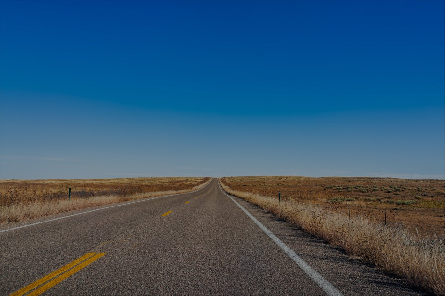 Open Kansas two-lane highway disappearing into the horizon, surrounded by grasslands and a clear blue sky.